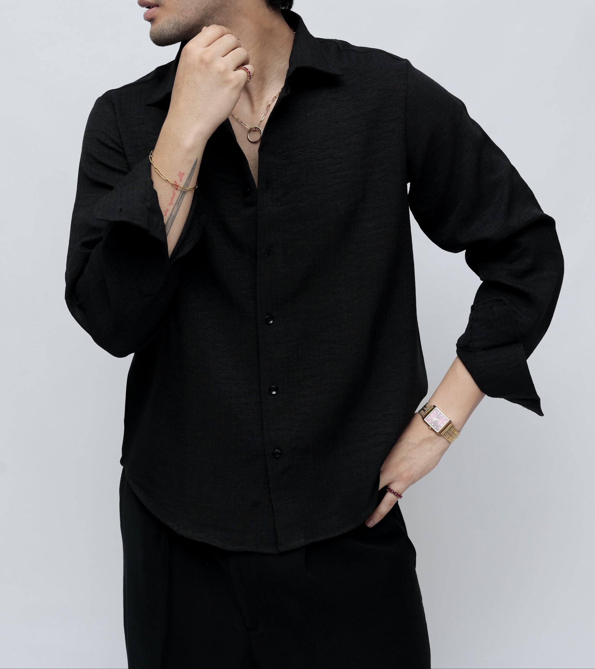 A man wearing a black long-sleeve button-up shirt and black trousers, posing against a light gray studio background.