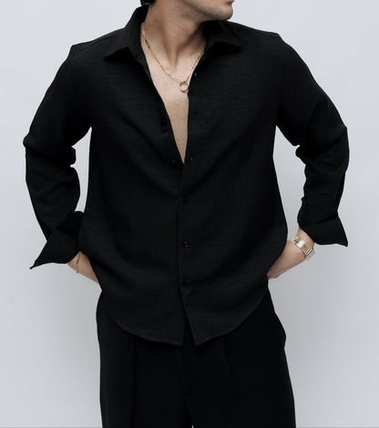 A man wearing a black long-sleeve button-up shirt and black trousers, standing against a light gray studio background.