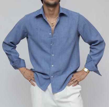 A man modeling a light blue long-sleeve button-up shirt and white trousers, standing against a plain light gray studio background.
