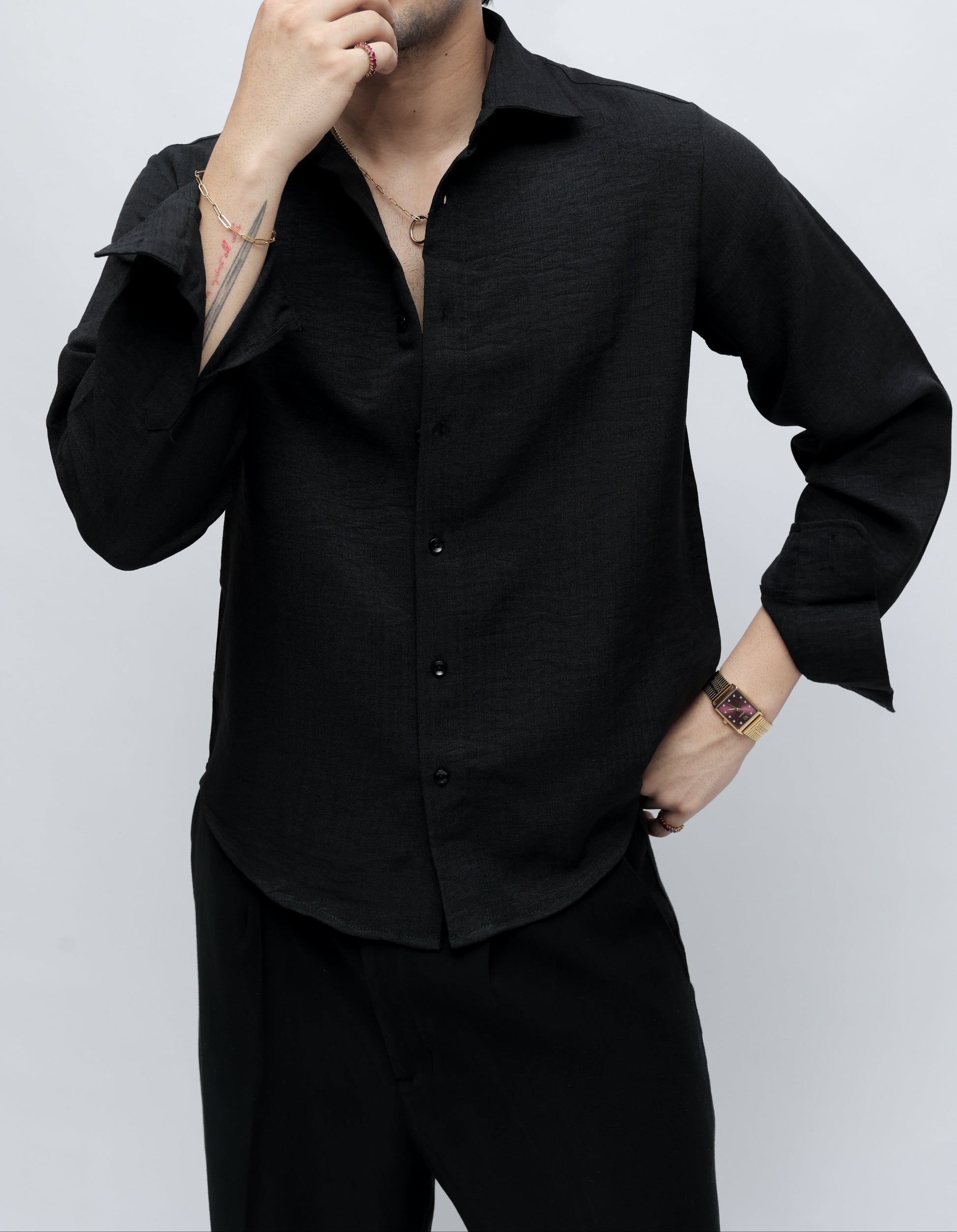 A man wearing a black long-sleeve button-up shirt and black trousers, posing against a light gray studio background.