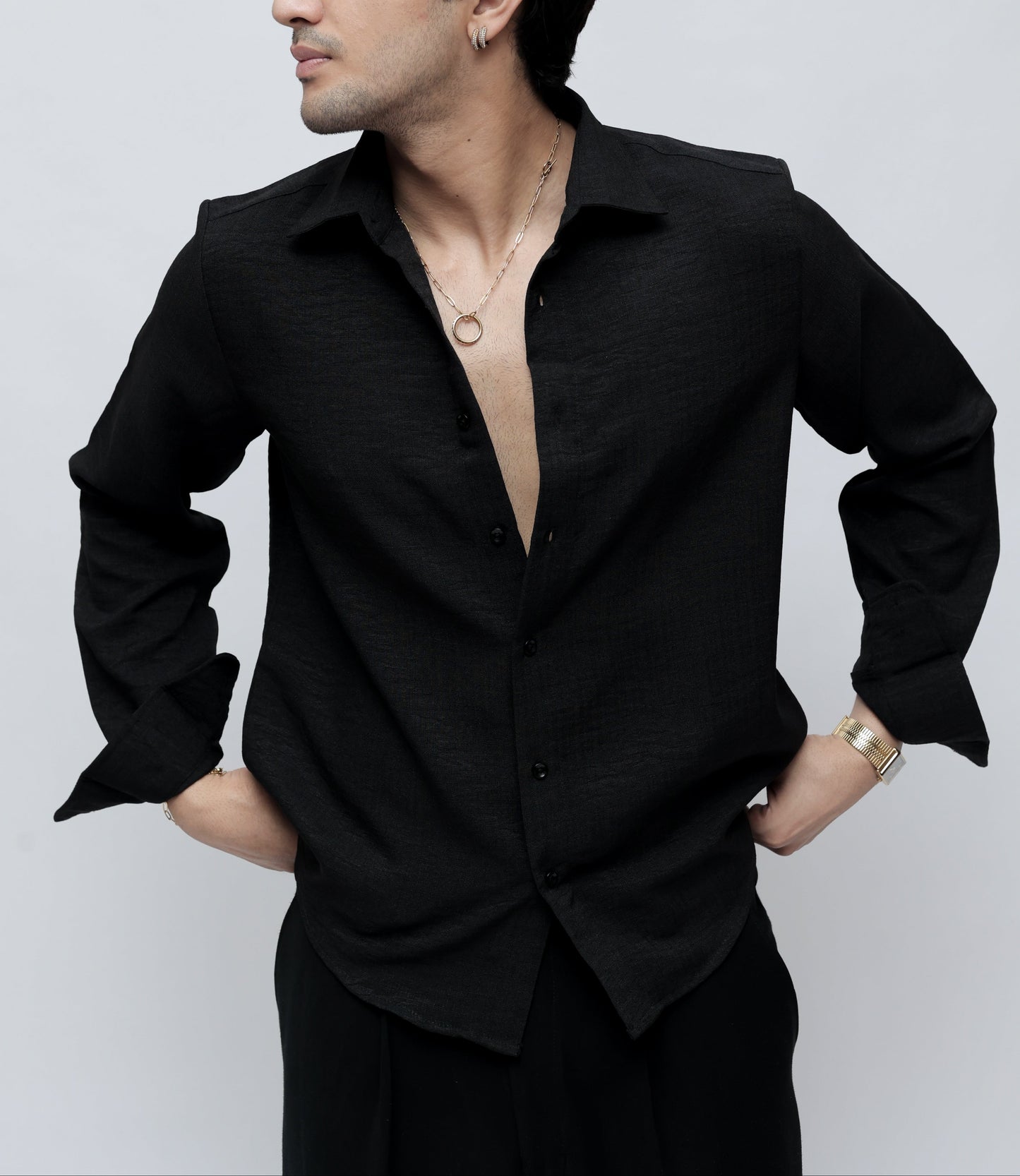 A man wearing a black long-sleeve button-up shirt with a relaxed, open-collar fit, posed against a light gray studio background.