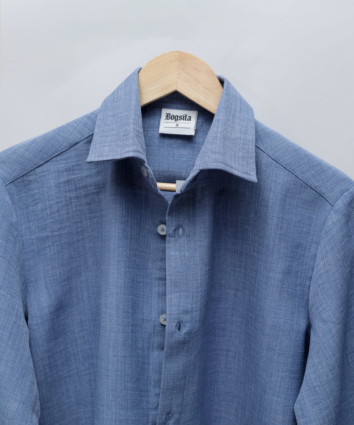 A blue textured long-sleeve button-down shirt hanging on a wooden hanger against a light gray background. The brand label inside the collar reads 'Bogula'.
