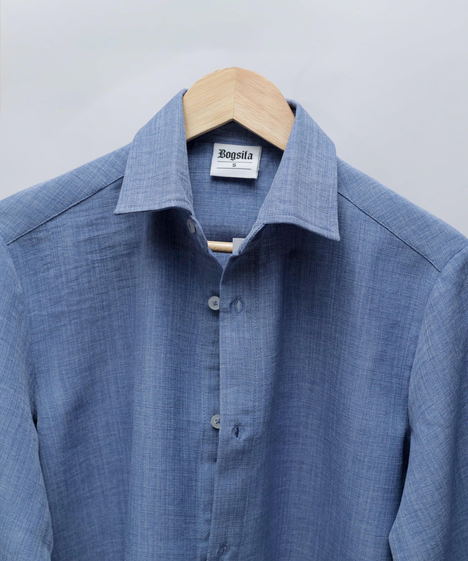 A blue textured long-sleeve button-down shirt hanging on a wooden hanger against a light gray background. The brand label inside the collar reads 'Bogula'.