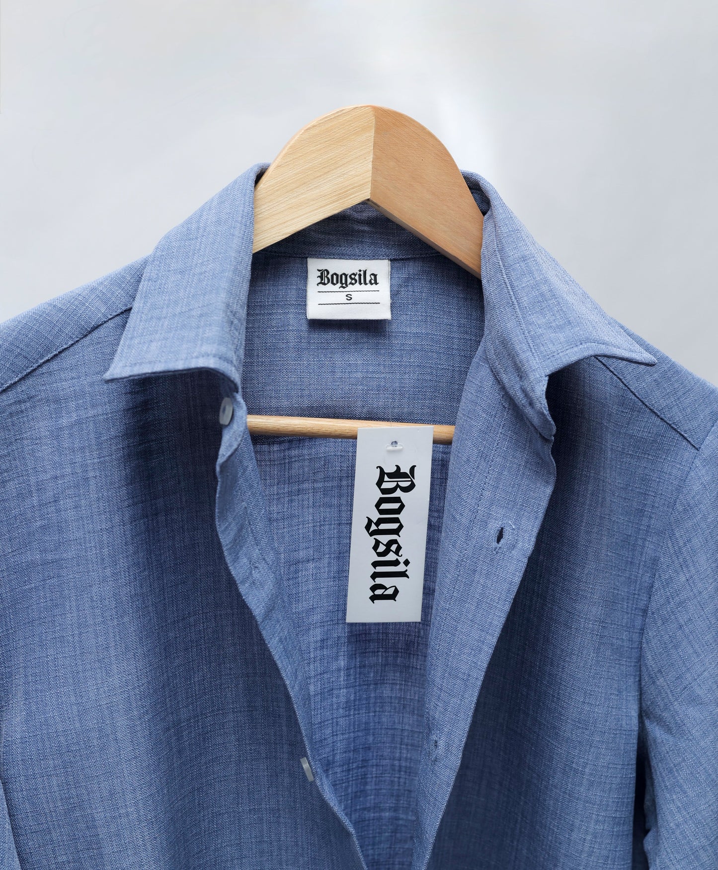 A blue textured linen button-down shirt hanging on a wooden hanger against a light gray background. The brand name 'Bogsla' is clearly visible on both the inner neck label and the hanging paper tag.