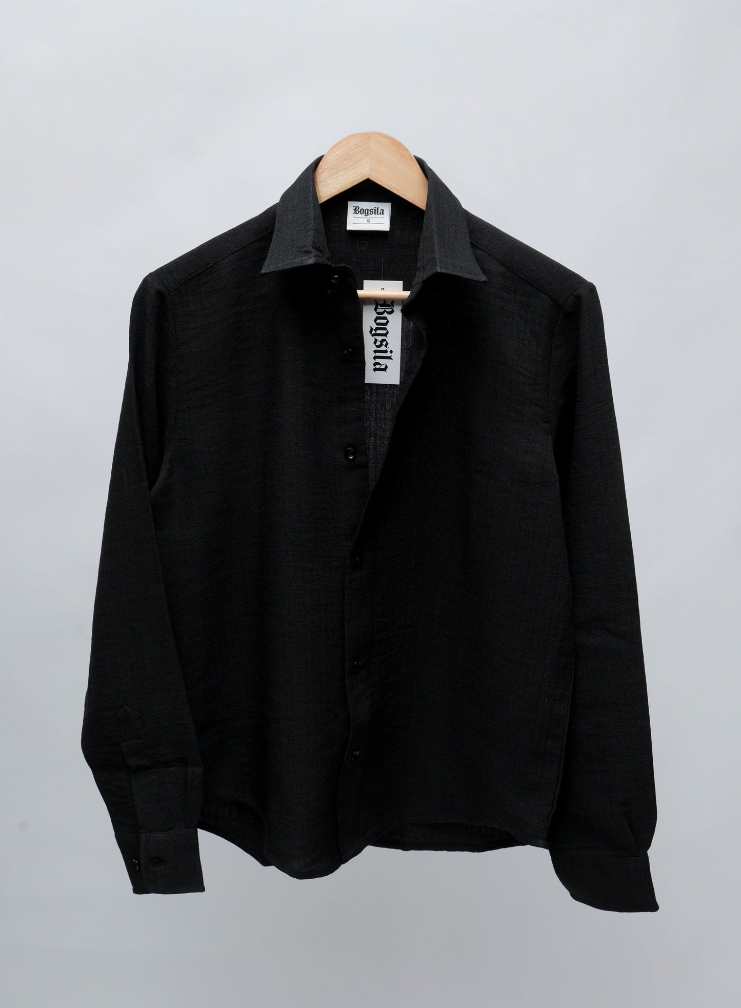 A black long-sleeve button-up shirt with a pointed collar, hanging on a wooden hanger against a light gray background.