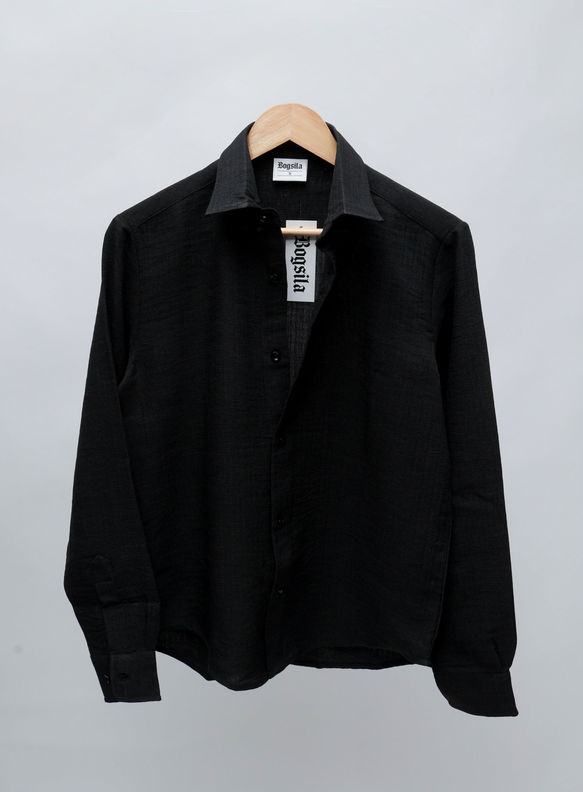 A black long-sleeve button-up shirt with a pointed collar, hanging on a wooden hanger against a light gray background.