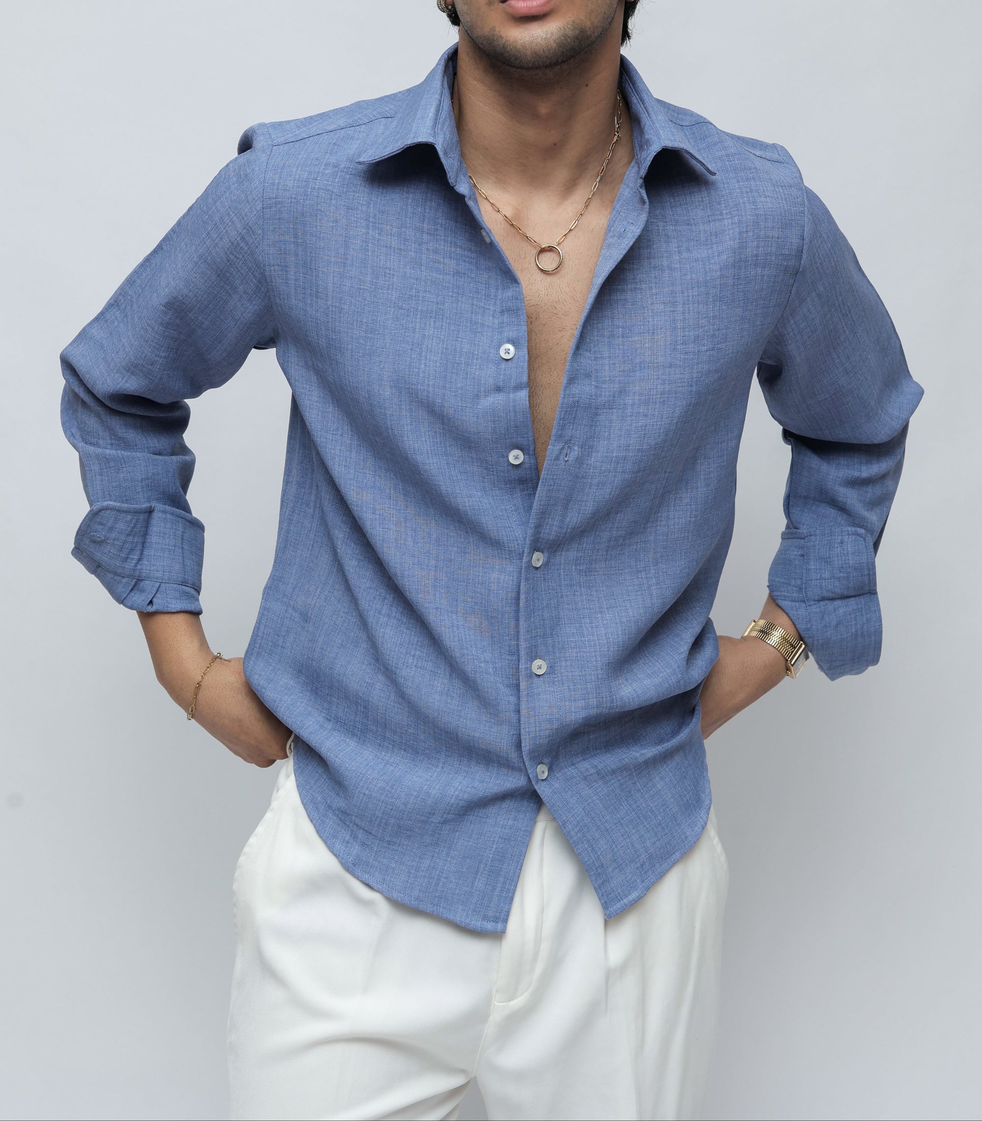 A man wearing a light blue long-sleeve linen button-up shirt and white trousers, standing against a light gray studio background.