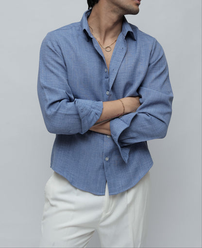A man wearing a light blue long-sleeve button-up shirt and white trousers, standing against a plain light gray studio background.