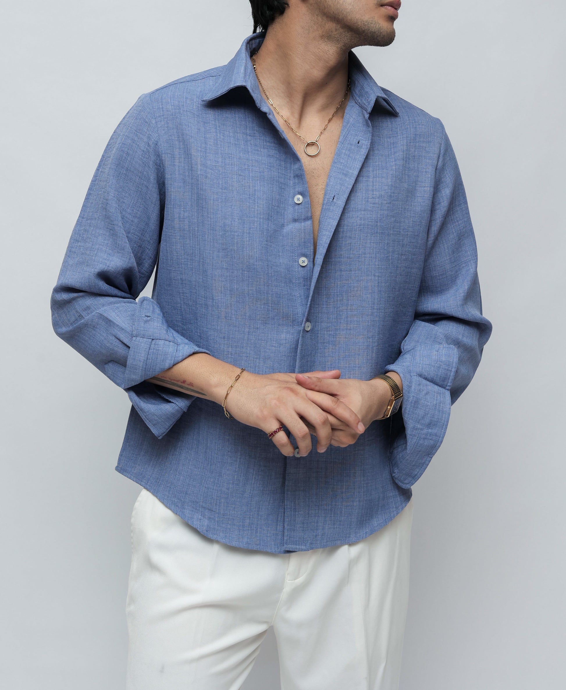A man wearing a light blue textured linen button-down shirt and white trousers, standing against a light gray studio background.