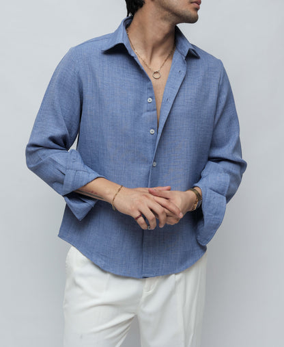 A man wearing a light blue textured linen button-down shirt and white trousers, standing against a light gray studio background.