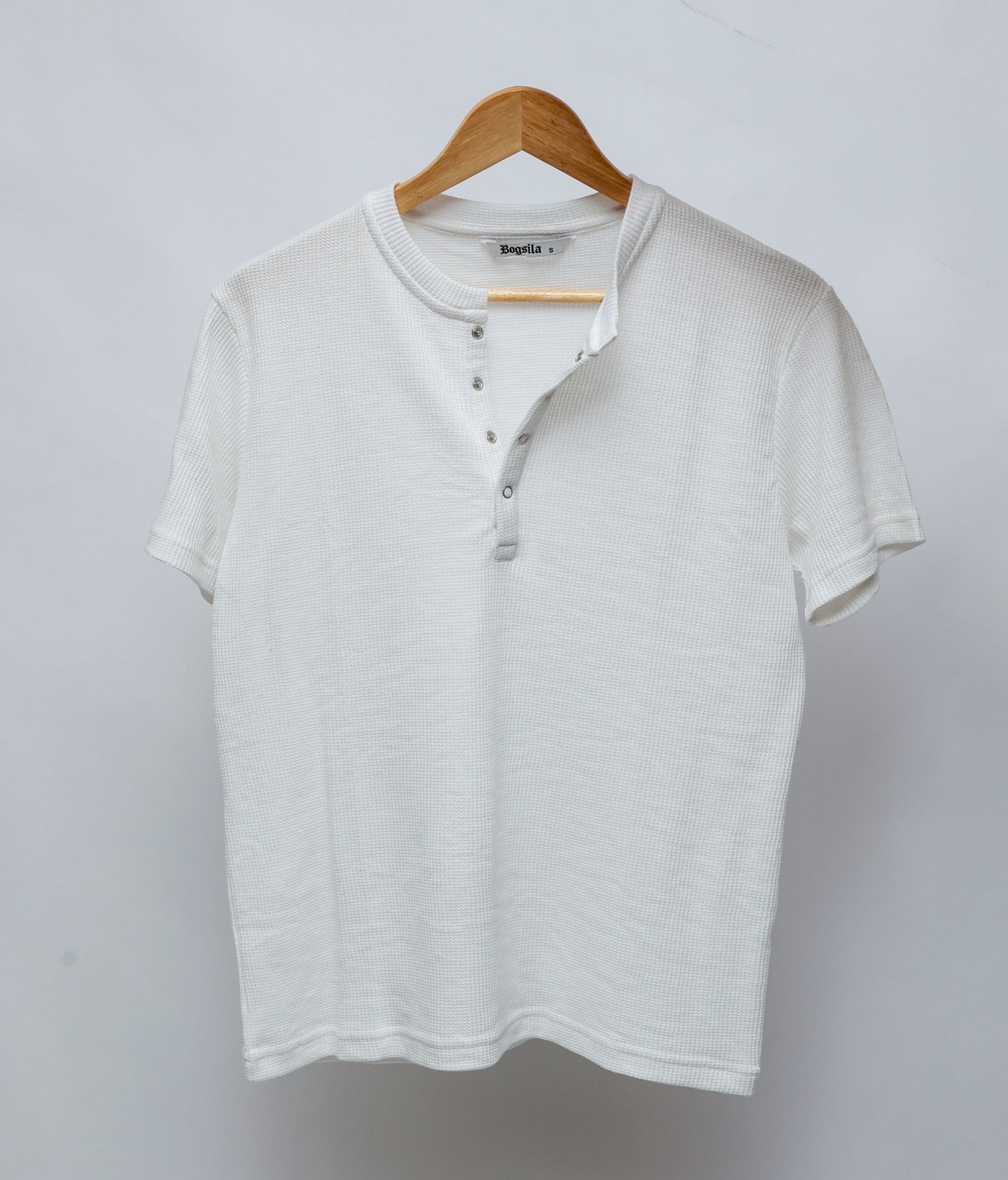 A white short-sleeve Henley T-shirt with a ribbed collar with four clip on button placket, hanging on a wooden hanger against a light gray background.