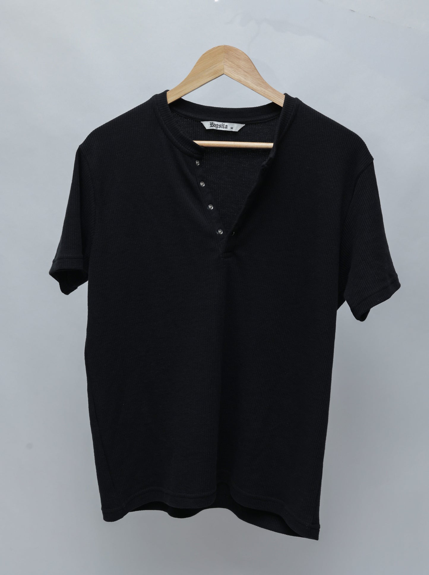 A Black short-sleeve Henley T-shirt with a ribbed collar and four-button placket, hanging on a wooden hanger against a light gray background.