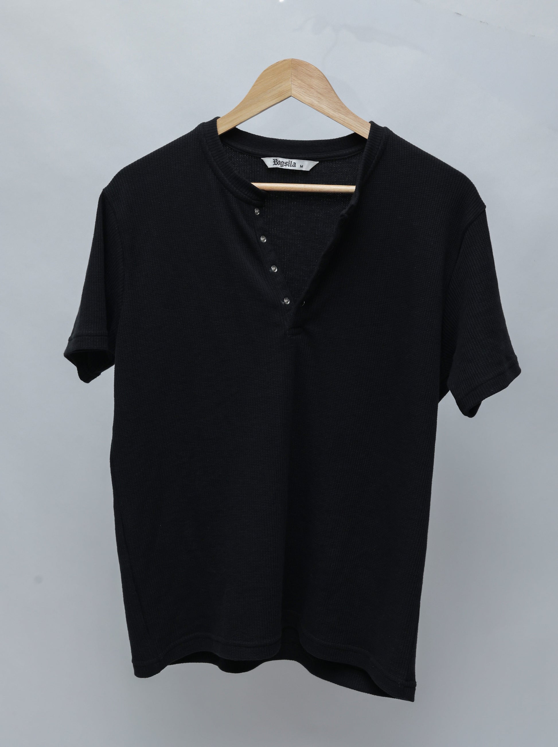 A Black short-sleeve Henley T-shirt with a ribbed collar and four-button placket, hanging on a wooden hanger against a light gray background.