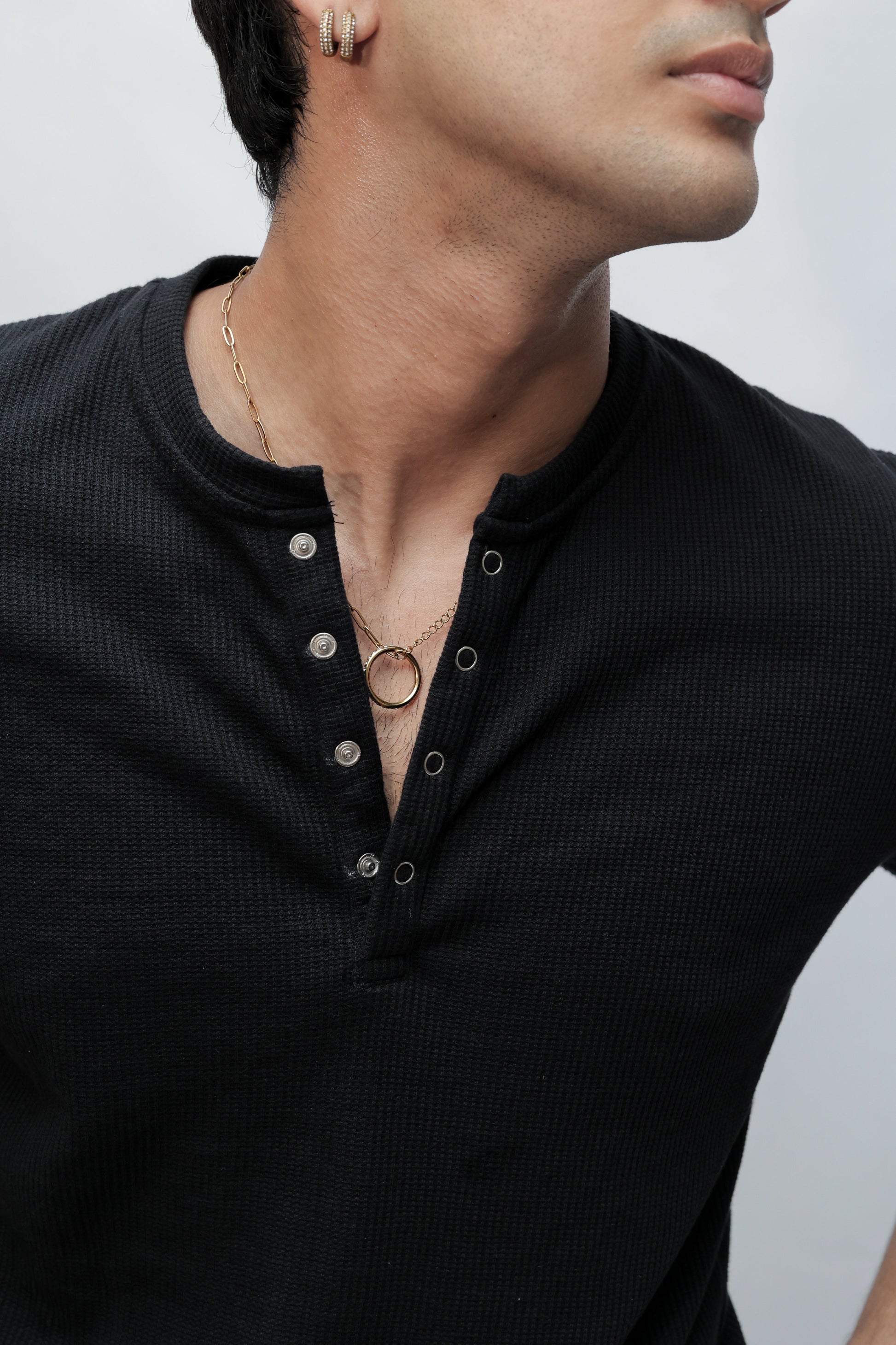 Close-up of a person wearing a black ribbed henley-style top with a split collar and metal snap details, paired with layered gold necklaces, against a light gray background.
