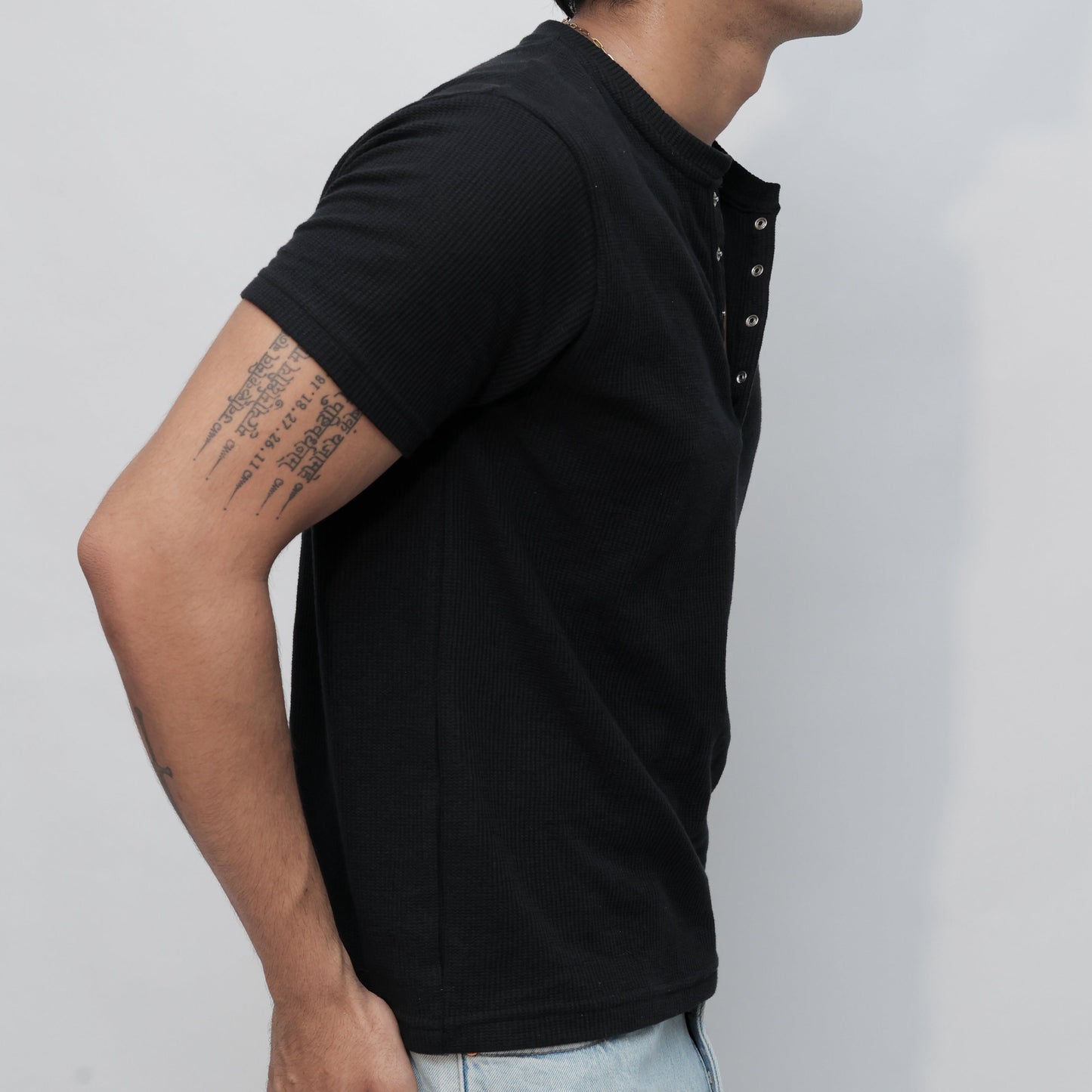 A man wearing a black short-sleeve t-shirt with a high collar and snap-button placket, paired with light blue jeans, shown from a side profile against a light gray background.