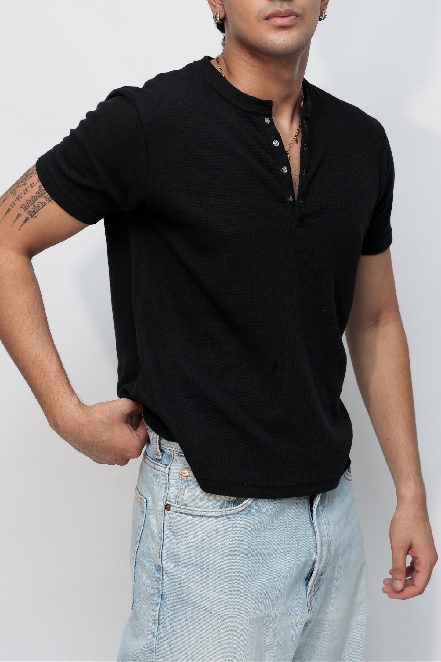 A man wearing a black short-sleeve polo shirt and light blue jeans, posed diagonally against a clean white studio background.