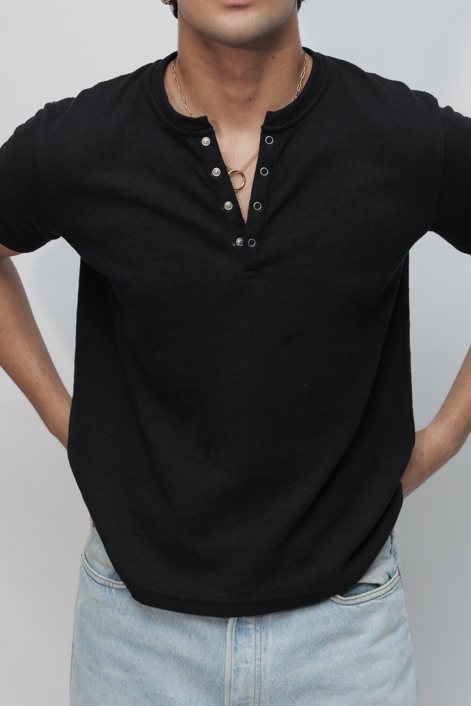 A man wearing a black short-sleeve henley shirt and light blue jeans, photographed from a high-angle perspective against a plain light gray studio background.