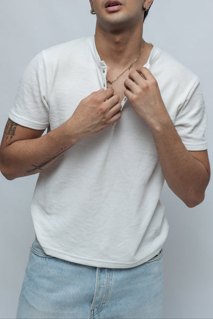 A young man wearing a white short-sleeve henley t-shirt and light blue jeans, looking up and adjusting the collar of the shirt against a plain light gray background.