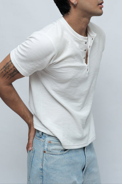 A man wearing a white short-sleeve henley shirt and light blue jeans, shown from a side profile against a plain light gray studio background.
