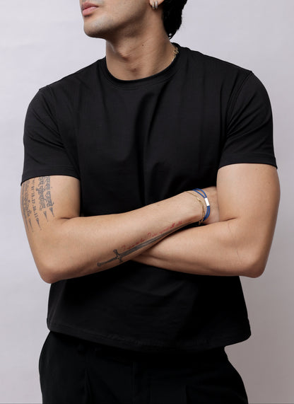 A man wearing a black t-shirt and black pants, lying down with his arms crossed, against a plain light gray background.